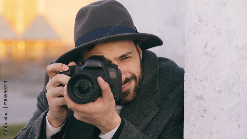 Closeup of young paparazzi man in hat photographing celebrities on camera while spy behind the wall Closeup of young paparazzi man in hat photographing celebrities on camera while spy behind the wall