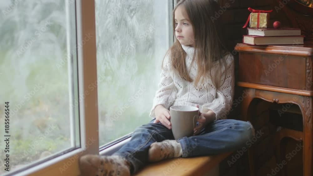 beautiful little girl with a mug of hot tea sits by the window, looks at the street and what she dreams about beautiful little girl with a mug of hot tea sits by the window, looks at the street and what she dreams about