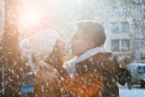 Cute lovely hipster couple with closed eyes in snowy winter city warming eachother on a bright day hugging each other and smiling. Tenderness  relationship concept. Snowfall effect.