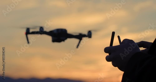 Close view of drone pilot's hands as he controls quadcopter hovering then flying off in the background. Aircraft and pilot silhouetted against sunset sky. Slow motion recorded at 60fps, 4K.