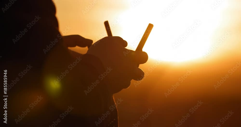 Close view of pilot's hands with remote control as he flies a drone at sunset. Strongly backlit with silhouette and lens flares. Hand-held real-time 4K.