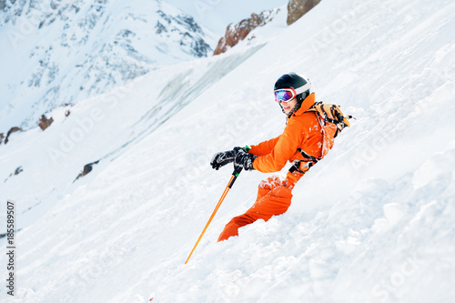 A skier in an orange overall and with a backpack is sitting happy in the snow after falling.