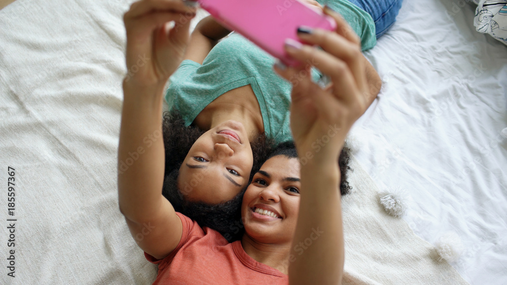 Top view of beautiful mixed race funny girlfriends making selfie portrait on bed in bedroom at home
