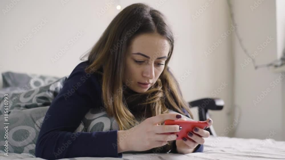 Cheerful young girl lying under blanket and typing on mobile phone in bed Cheerful young girl lying under blanket and typing on mobile phone in bed