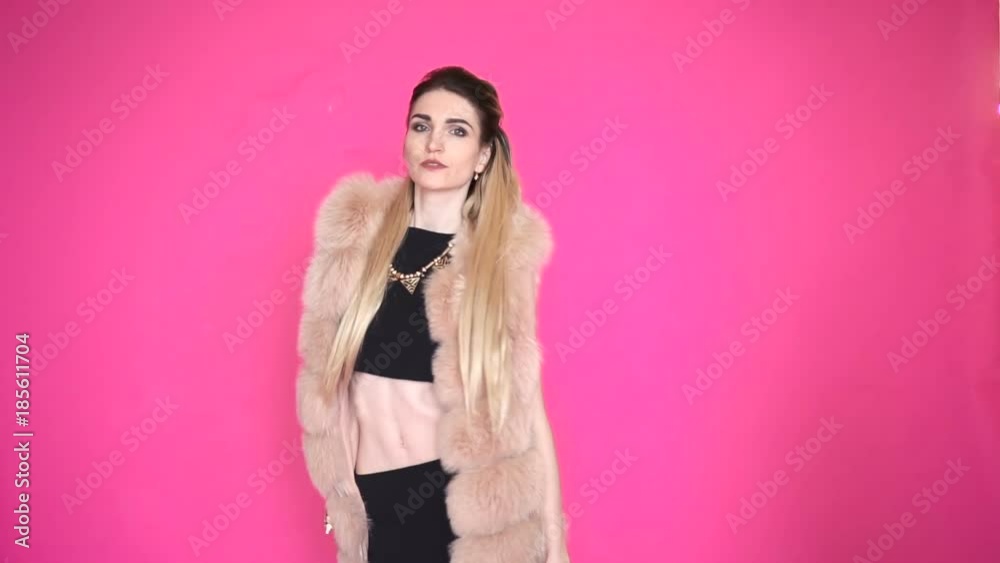 Beautiful girl with long dyed hair in a beautiful fur vest, is dancing on pink background.