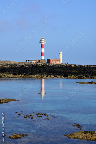 Leuchtturm mit weiß und roten Streifen und sein Spiegelbild im ruhigen Wasser.

