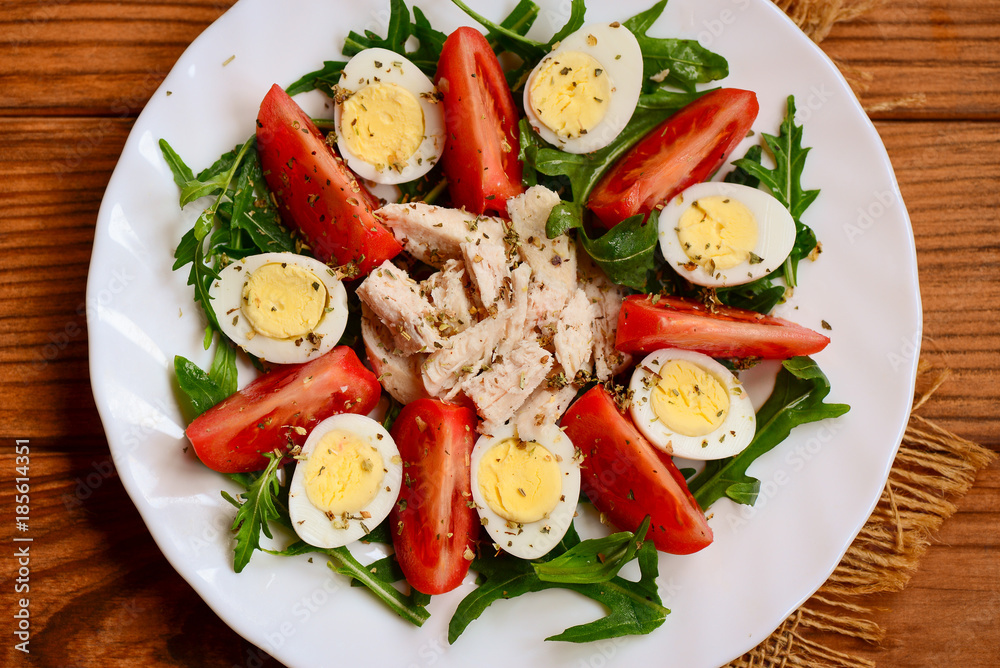 Fresh vegetable salad with boiled chicken breast and quail eggs. Salad with fresh tomatoes, rucola, quail eggs, chicken breast and spices on a serving plate and a wooden background. Closeup. Top view Fresh vegetable salad with boiled chicken breast and quail eggs. Salad with fresh tomatoes, rucola, quail eggs, chicken breast and spices on a serving plate and a wooden background. Closeup. Top view