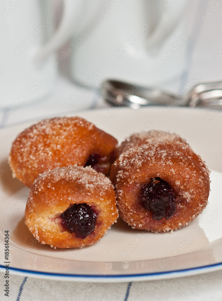 Jelly filled brioche donut hole /  three freshly baked, warm Jelly filled brioche donut holes on a white, round plate with a blue rim on a white table cloth with thin blue stripes 