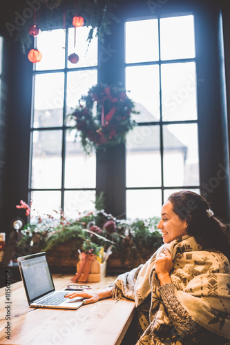 beautiful young girl uses laptop technology,types text looking at monitor in cafe by window at wooden table,in winter decorated with Christmas decor.Dressed in gray knitted wool sweater and glasses