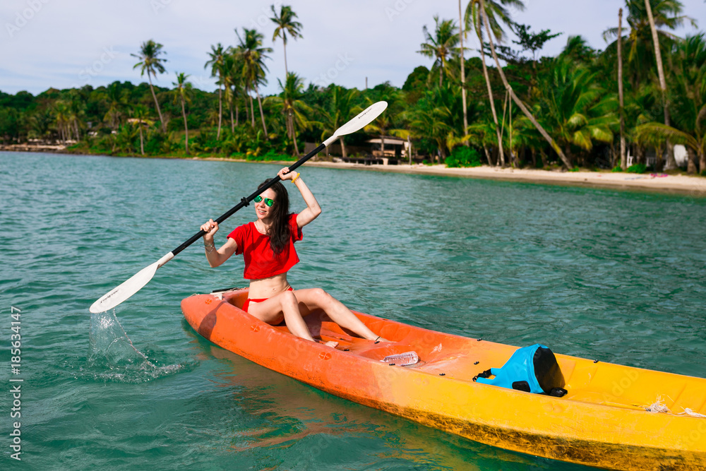 Brunette young woman girl in red bikini paddling  on red, orange, yellow kayak in the blue water. Asia. Thailand