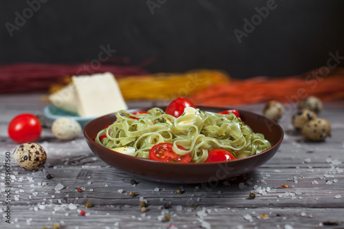 pasta from green spinach with mozzarella, greens and French mustard, quail eggs and cherry tomatoes on a plate, bell pepper and large sea salt on a gray wooden table on a dark background