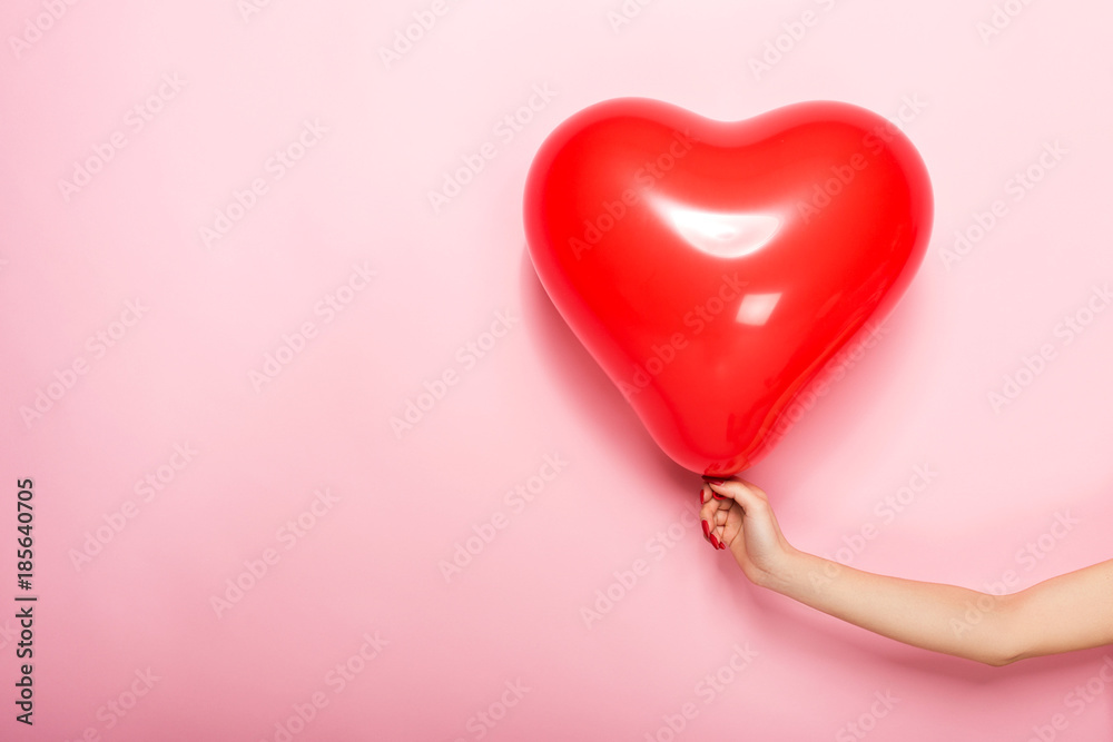 Fototapeta premium Female hand holding a red ball in the shape of a heart, isolated on a pink background.