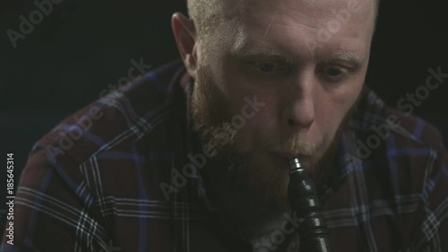 closeup bearded serious guy man smoking hookah, fume shisha and looking perceptively at the camera in the dark room. photo