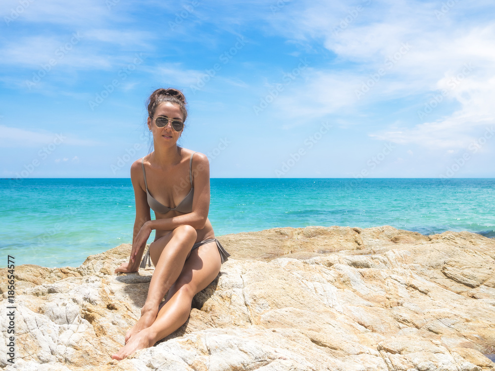 Sun-tanned young girl in gray bikini sitting on the stony shore of the sea. View from the front