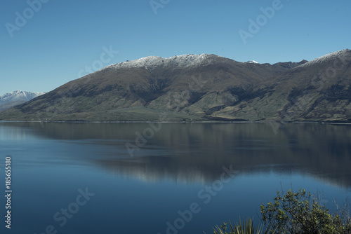 Paisaje de monta  as frente a un gran lago donde se reflejan. Cielo azul despejado.