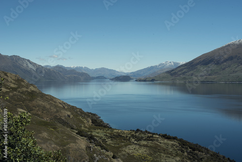 Paisaje de montañas frente a un gran lago donde se reflejan. Cielo azul despejado.