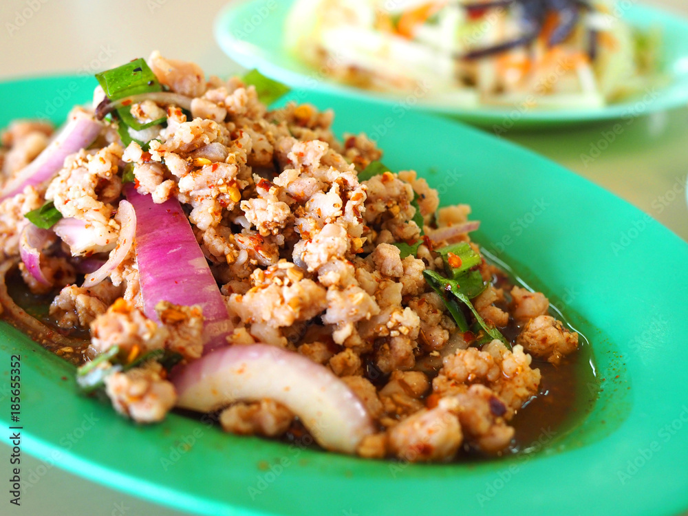 Close up Thai local traditional Isaan Northest Larb Moo, raw semi stir fried spicy minced pork with shallot, spring onion, and herb, on green oval plastic plate, with blurred Som Tam, papaya salad Close up Thai local traditional Isaan Northest Larb Moo, raw semi stir fried spicy minced pork with shallot, spring onion, and herb, on green oval plastic plate, with blurred Som Tam, papaya salad