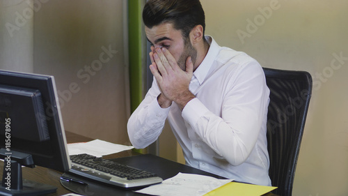 Tired bored young businessman sitting at his desk in front of his computer with his chin resting on his hand, sleepy photo