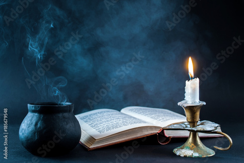 Candle in candlestick burning and smoky pot and old open book on dark and smoked background. Halloween concept