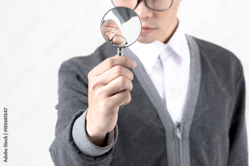 Nerd studying hard. Young Asian nerd man wearing glasses holding magnify glass standing isolated on white background. Black hair, grey sweater, white shirt. Education concept. Nerd studying hard. Young Asian nerd man wearing glasses holding magnify glass standing isolated on white background. Black hair, grey sweater, white shirt. Education concept.