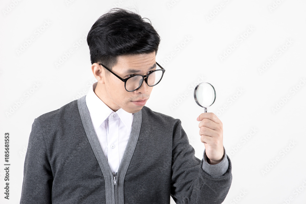 Nerd studying hard. Young Asian nerd man wearing glasses holding magnify glass standing isolated on white background. Black hair, grey sweater, white shirt. Education concept. Nerd studying hard. Young Asian nerd man wearing glasses holding magnify glass standing isolated on white background. Black hair, grey sweater, white shirt. Education concept.