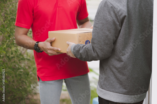 Young logistic career concept. Happy delivery man giving his package to customer at home. Taken in real house. Asian chinese fit man in red polo shirt and jeans with red hat in his early twenties.