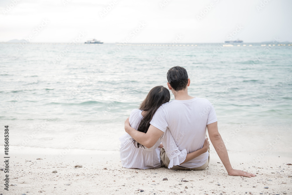 Couple holding each other on beach. Young happy interracial couple on beach holding each other sitting down. Asian woman, Caucasian man. Young mixed race romance concept.