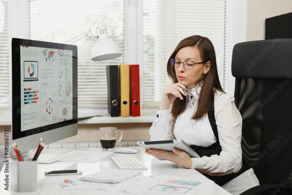 Beautiful engrossed brown-hair business woman in suit and glasses sitting at the desk with cup of coffee, mobile phone, working at computer with modern monitor with document and tablet in light office Beautiful engrossed brown-hair business woman in suit and glasses sitting at the desk with cup of coffee, mobile phone, working at computer with modern monitor with document and tablet in light office