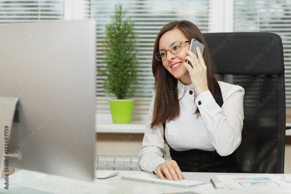 Beautiful smiling business woman in suit and glasses sitting at the desk, working at contemporary computer with documents in light office, talking on mobile phone, conducting pleasant conversation Beautiful smiling business woman in suit and glasses sitting at the desk, working at contemporary computer with documents in light office, talking on mobile phone, conducting pleasant conversation