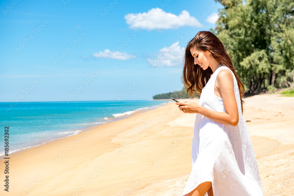 Beautiful young single white woman on beach. Using mobile phone calling her friends, wearing white dress. Connected world concept.