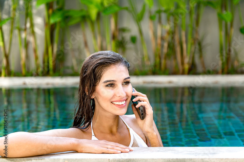 Beautiful young single white woman in pool. Talking on mobile phone, wearing white bikini. Connected world concept.