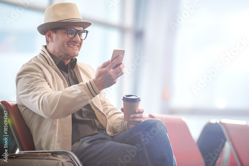Surfing internet. Joyful adult man in hat and glasses is waiting for flight at airport lounge. He is using mobile phone with smile while drinking hot coffee. Copy space in the right side