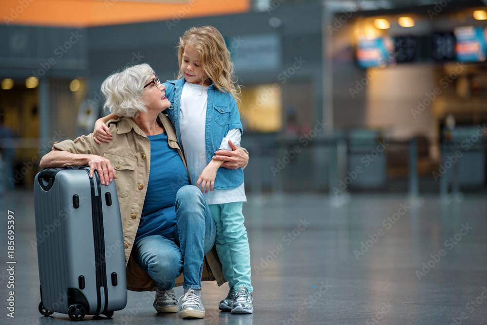Full length of happy elegant gray-haired grandmother is squatting and resting on suitcase in waiting hall while hugging her small grandchild. They are looking at each other with smile. Copy space
