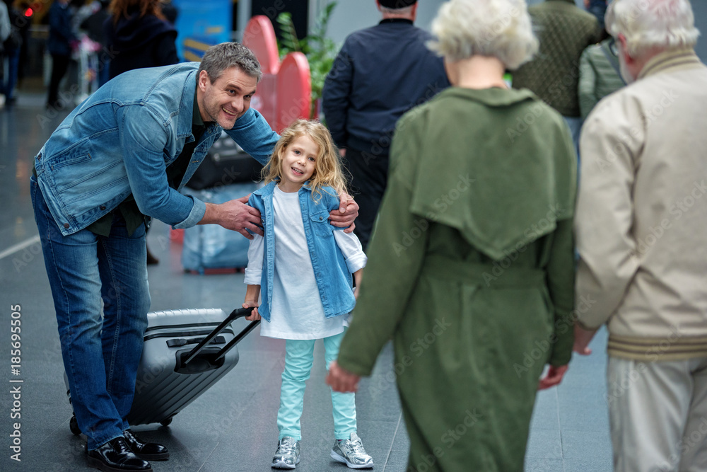 Fototapeta premium Say goodbye. Grandparents are accompanying adult son and his daughter to airport. They are standing back to camera. Focus on positive man hugging little girl with suitcase