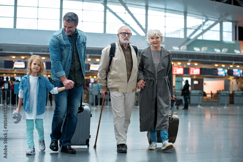 Full length of delighted old grandparents are going with their son and granddaughter at airport terminal to check-in on their flight. They are carrying suitcases and expressing happiness