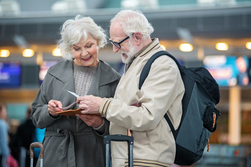 What time our plane. Involved gray-haired couple is standing at airport terminal and holding flight tickets. They are checking travel documents while discussing together with smile