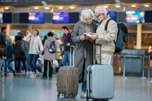 Checking trip details. Full length of serious senior wife and husband are standing with suitcases at international airport and looking at flight tickets with concentration. Copy space in left side