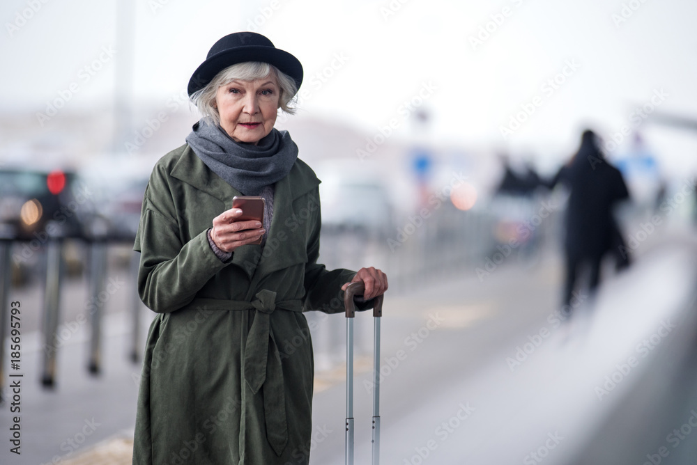 Lost in thoughts. Portrait of elegant senior lady in hat is standing outdoors with suitcase. She is holding modern mobile phone and looking aside thoughtfully. Copy space in the right side Lost in thoughts. Portrait of elegant senior lady in hat is standing outdoors with suitcase. She is holding modern mobile phone and looking aside thoughtfully. Copy space in the right side