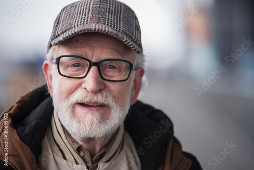 Good day. Close-up portrait of cheerful gray-haired bearded gentleman in glasses is standing outdoors. He is looking at camera with joy. Copy space in the right side