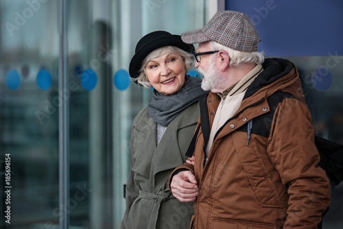 Feeling excitement. Elegant old woman is going under the handle with her husband. She is looking at him with happy smile while exiting from airport building. Copy space in the left side