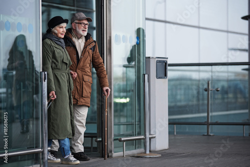 Finally at home. Full length of positive elegant old man and woman are going out of doors from terminal. Male is exiting with cane while female is carrying her suitcase. Copy space in the right side © Yakobchuk Olena
