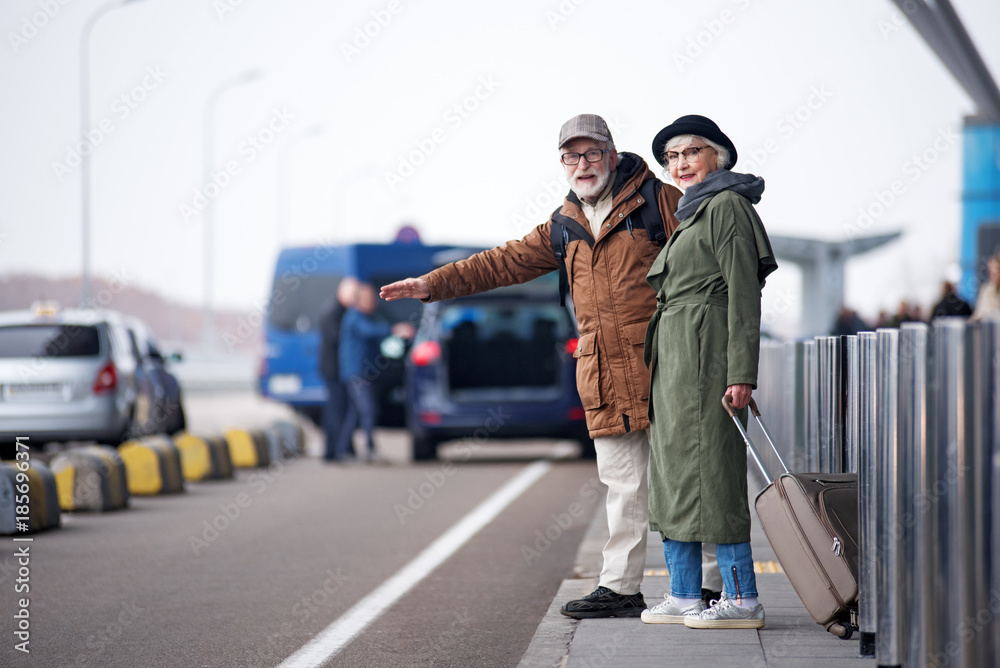 Attending new town. Full length of optimistic old man and woman are standing near road and trying to catch taxi. Gentleman is rising hand while lady is holding suitcase. Copy space on the left side Attending new town. Full length of optimistic old man and woman are standing near road and trying to catch taxi. Gentleman is rising hand while lady is holding suitcase. Copy space on the left side