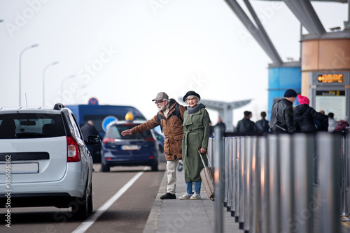 Hitchhiking. Full length of stylish senior couple is standing near road and trying to catch transport. Gentleman is rising hand while lady is looking at camera and holding her luggage confidently