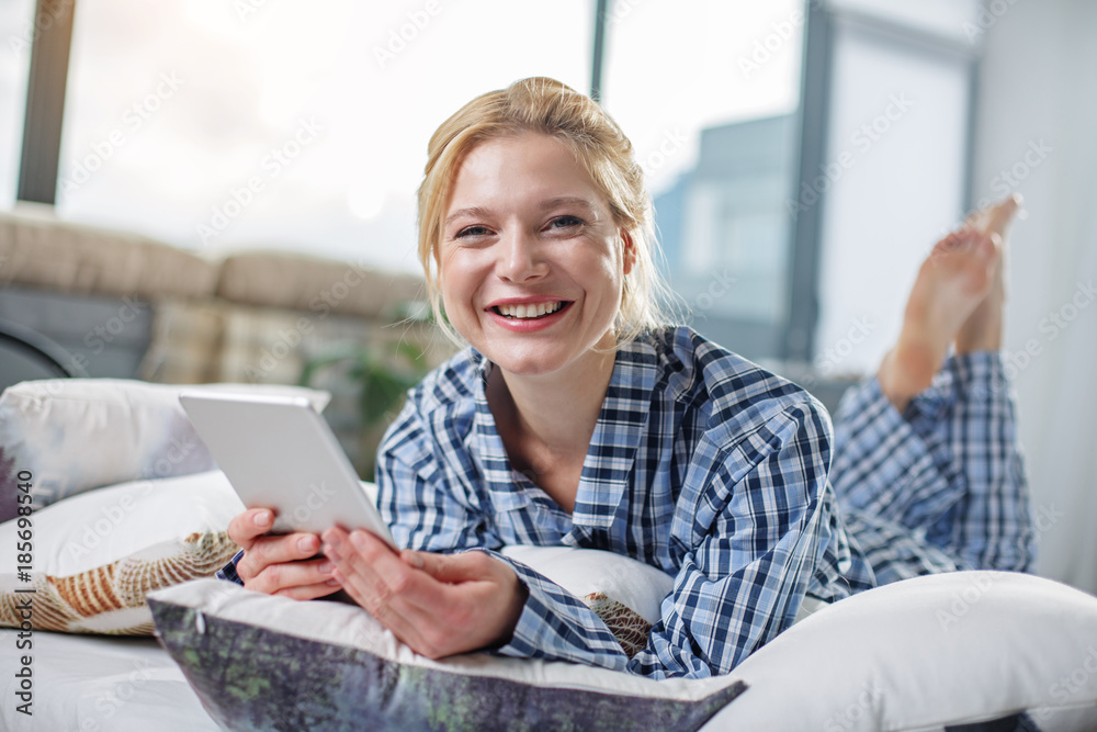 Portrait of laughing lady lying on home bed with gadget in hands. She is looking at camera with happiness Portrait of laughing lady lying on home bed with gadget in hands. She is looking at camera with happiness