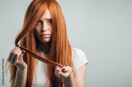 Sad redhead girl holding her damaged hair looking at camera