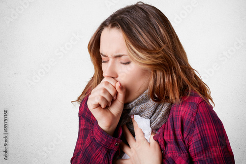 Fotografie, Obraz Studio shot of beautiful young female model has bad cough, uses tissue, wears scarf on neck, feels unwell and unhealthy, isolated over white concrete background