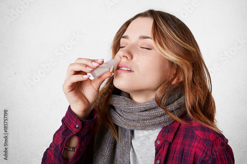 Sick pretty woman has blocked nose, dripps nasal drops, suffers from running nose, isolated over white concrete background. Allergic young female has nasal congestion. Healthcare concept