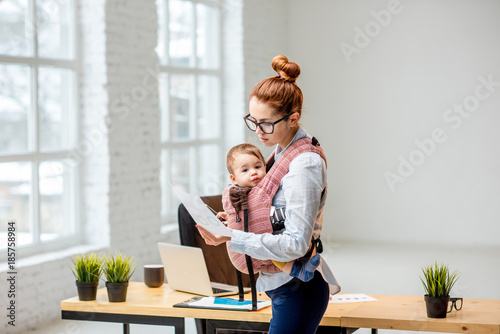 Young multitasking businesswoman standing with her baby son during the work at the office photo