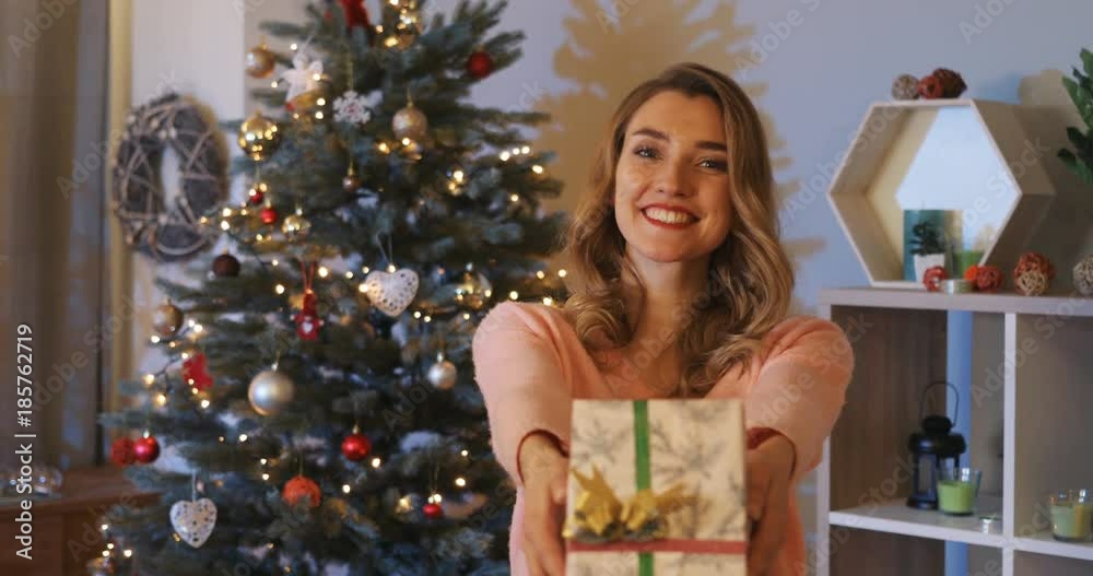 Pretty young smiling woman with curly hair giving present on background of beautiful Christmas tree with decorations in room of big house.