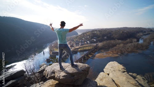 Young man stands on a rock and raises his arms at Maryland Heights while looking at the view of Harpers Ferry National Park and the Potomac and Shenandoah Rivers. photo